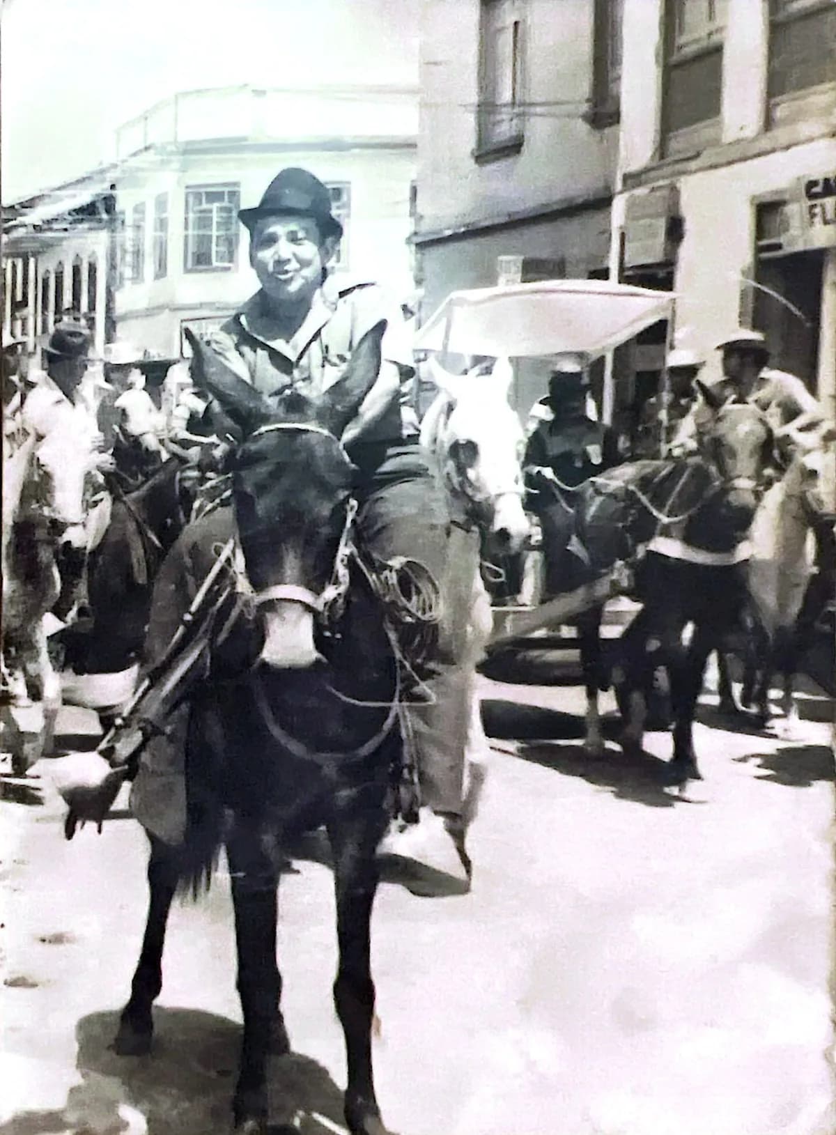 Fotografía antigua en blanco y negro: un arriero a caballo en la calle de un pueblo, entre otras bestias de carga