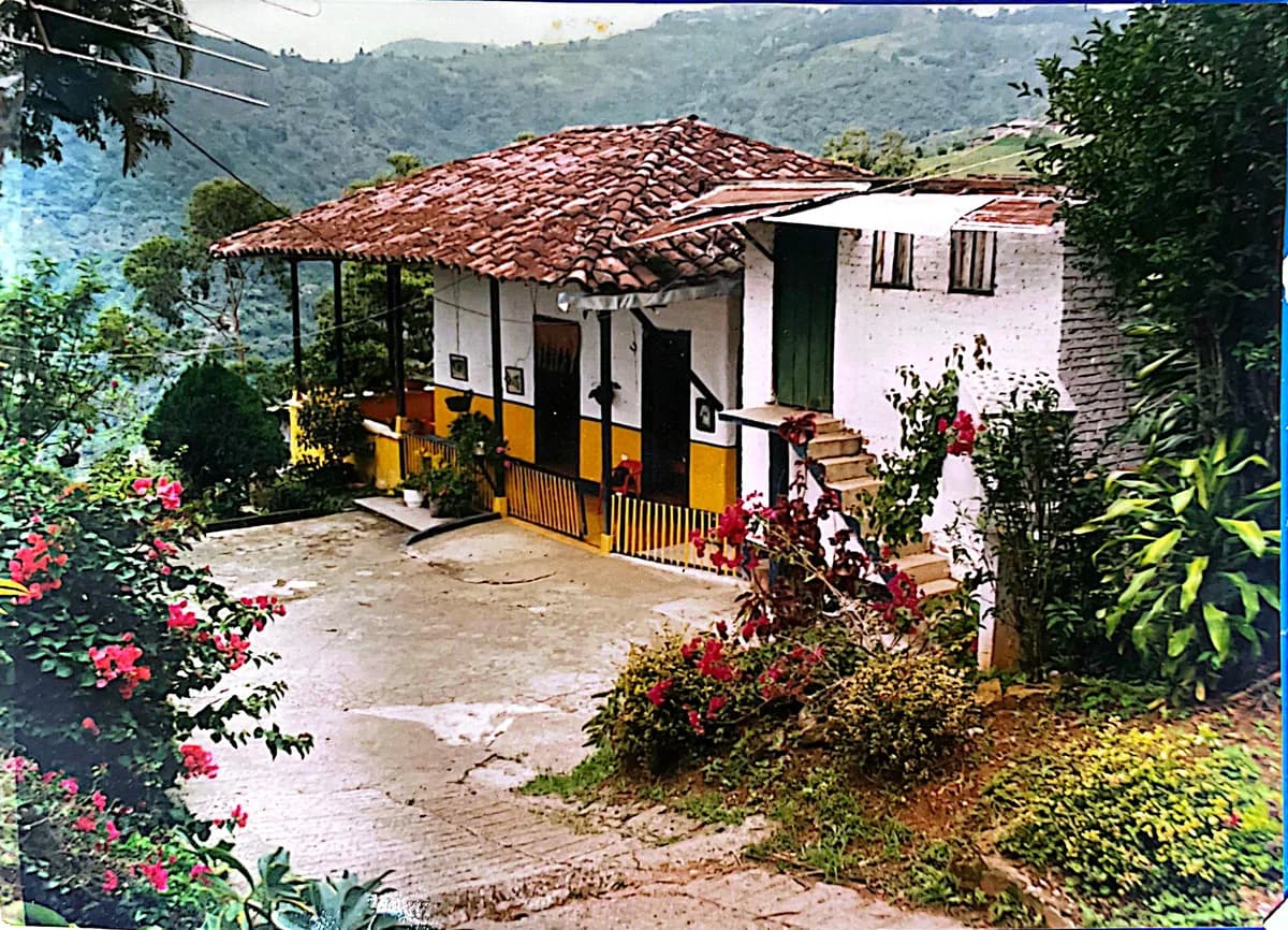 La casa de la finca: paredes blancas, corredor de madera pintado, tejado de barro y flores alrededor