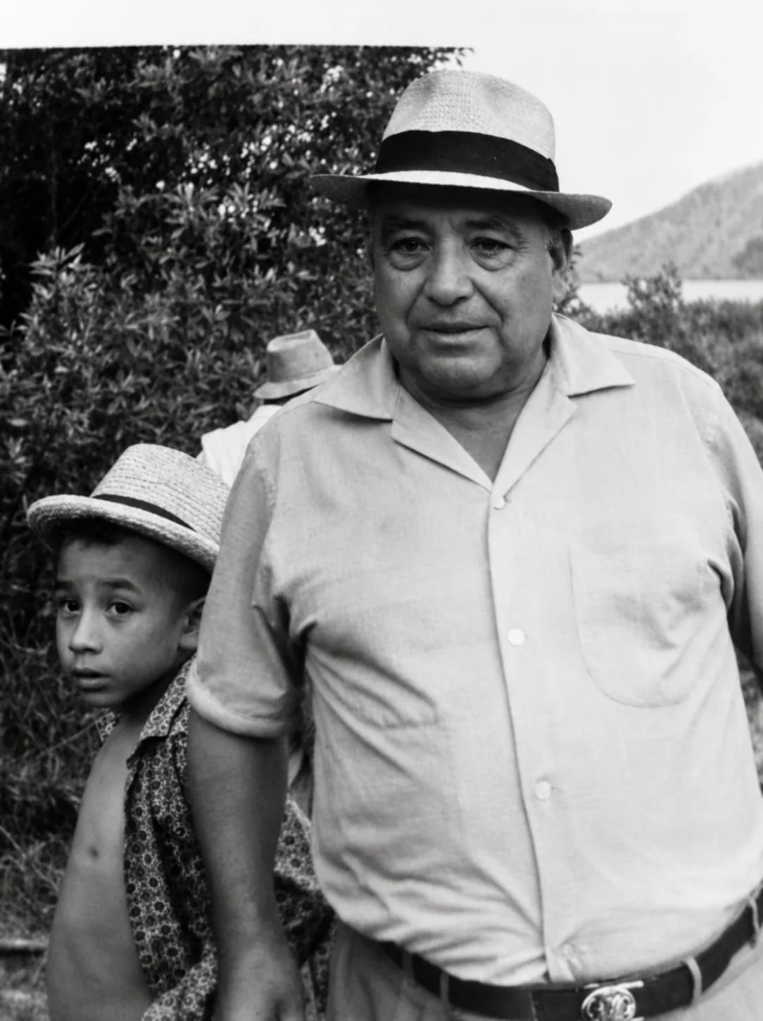 Fotografía antigua en blanco y negro: un hombre mayor con sombrero de paja, con un niño también con sombrero a su lado, entre la vegetación de la finca y con la montaña al fondo